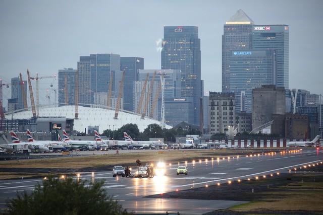 A photograph of the runway at London City Airport, taken from a distance at dawn. A small group of protesters can be seen surrounded by security vehicles. Grounded planes are lined up behind the runway. The skyscrapers of London's financial district rise above the scene.