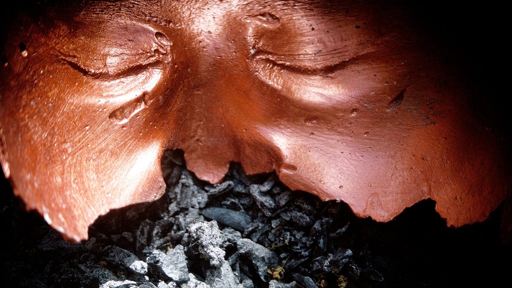A still from Maya Campbell's performance artwork entitled 'Adding A Face'. The still shows a close-up of a bronze-coloured mask fragment lying on ashes.
