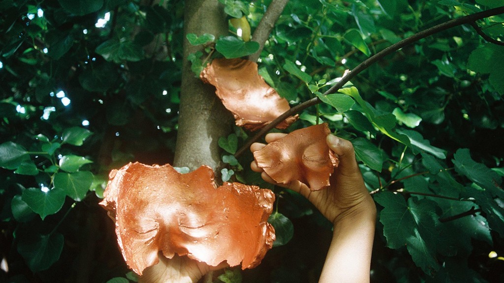 A still from Maya Campbell's performance artwork entitled 'Adding A Face'. The artist's hands hold bronze fragments of mask up against a background of foliage. One mask fragment is nestled in the branches of a tree.